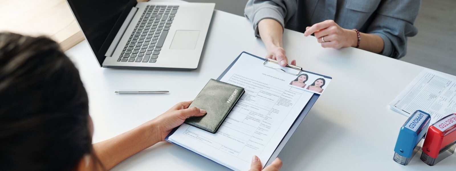 Two women passing a clipboard between then with a visa application form and passport on top.